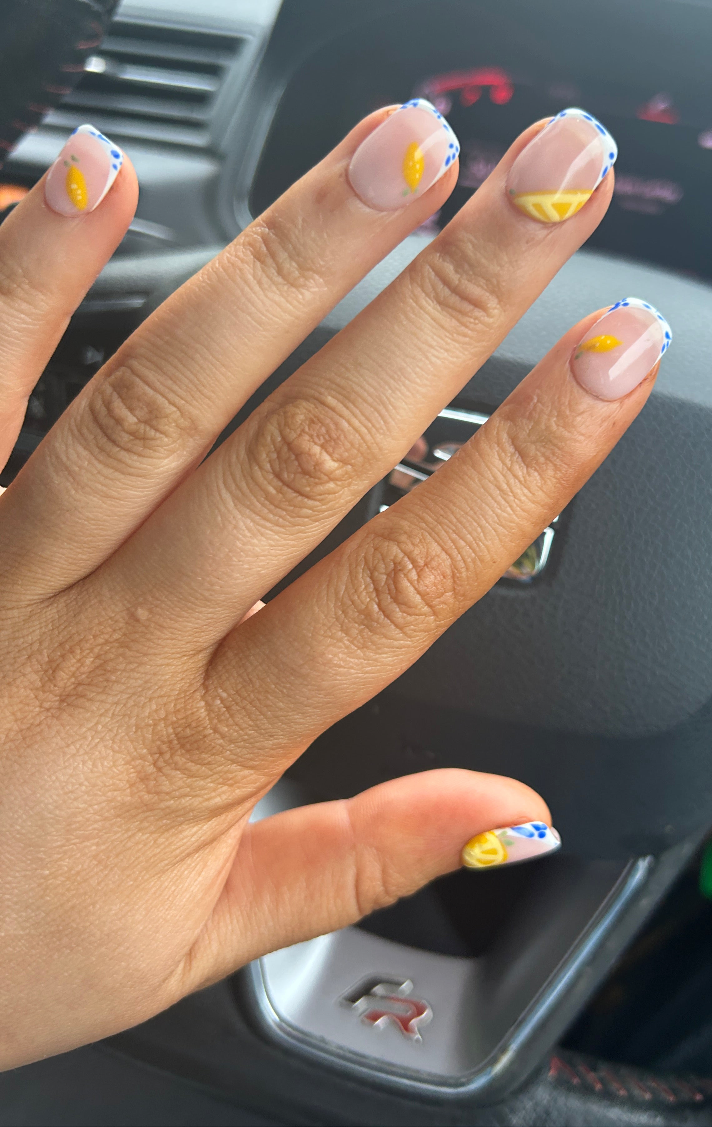 A hand with false nails featuring a nude base with French tips and subtle lemon designs, wearing a silver ring.