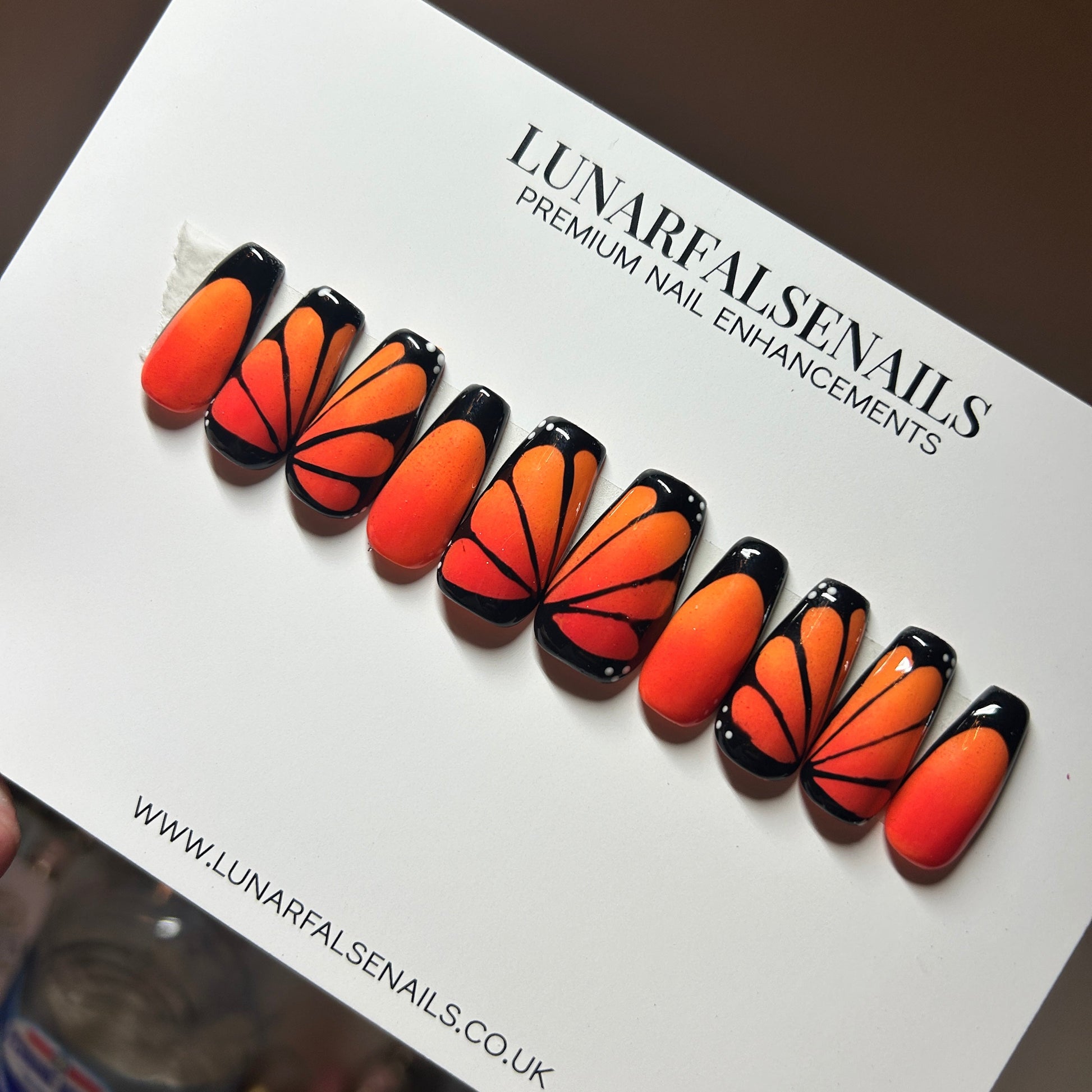 Butterfly-shaped orange and black nail enhancements on a LunarFalseNails branded card.